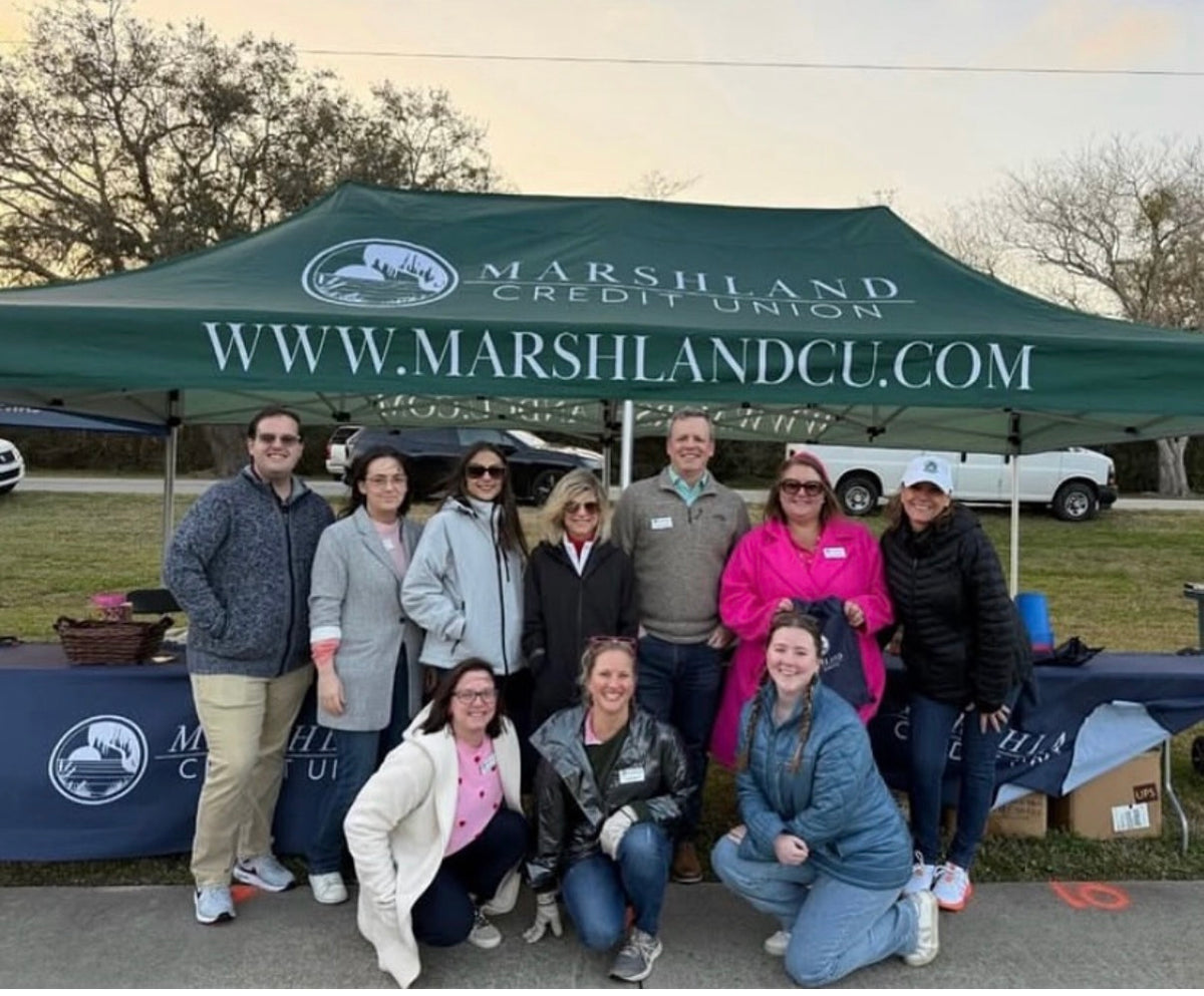 A group of nine people posing outdoors under a green Marshland Credit Union pop-up canopy tent, with a branded table in the foreground and trees and a vehicle in the background.