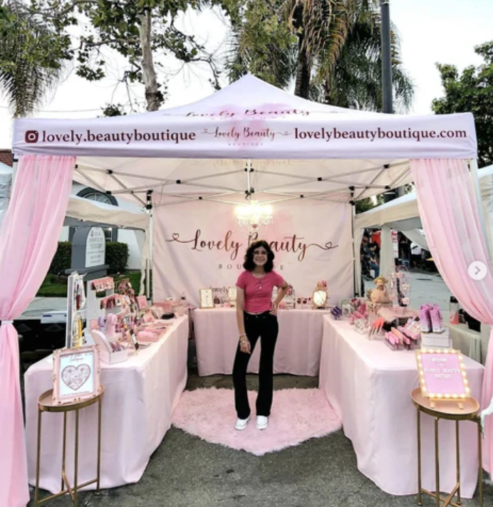 A woman stands behind a styled pop-up boutique stall at an outdoor market. The stall features a white canopy with pink draped curtains, a pink-and-white branded backdrop reading 'Lovely Beauty Boutique,' and display tables covered in pink cosmetics and beauty products.