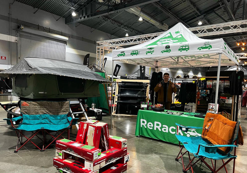 A ReRack booth at an outdoor equipment show, featuring a green branded table, a white canopy with green logos, and a man standing behind it. In the foreground, a green rooftop tent is mounted on a folding chair next to teal camping chairs and stacked product boxes.