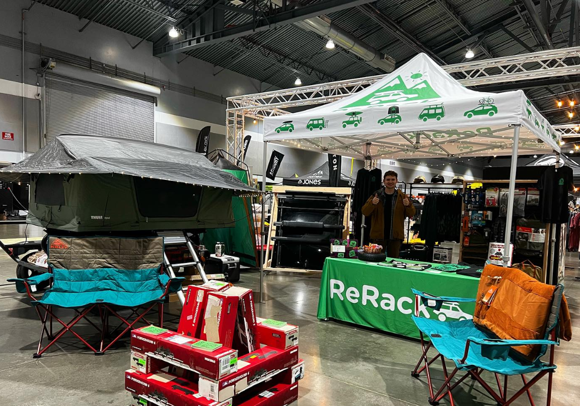 A ReRack brand pop-up canopy tent is set up at an outdoor gear exhibition or trade show, with a man standing behind the green branded table. Other camping gear, including a rooftop tent and folding chairs, are displayed around it in a large indoor hall.