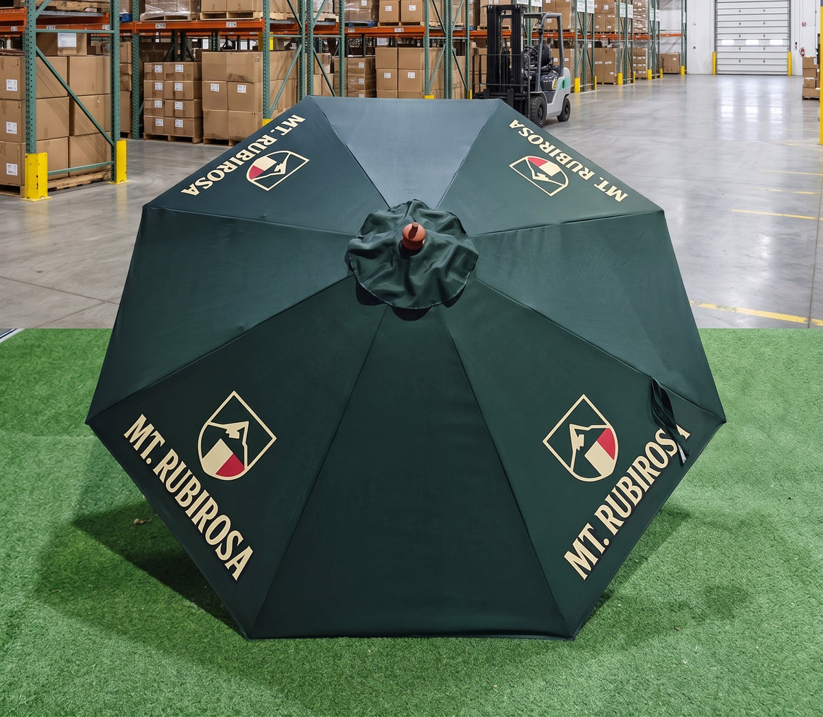 A dark green patio umbrella with gold 'MT. RUBIROS A' branding and logos, displayed open on artificial green turf inside a warehouse.