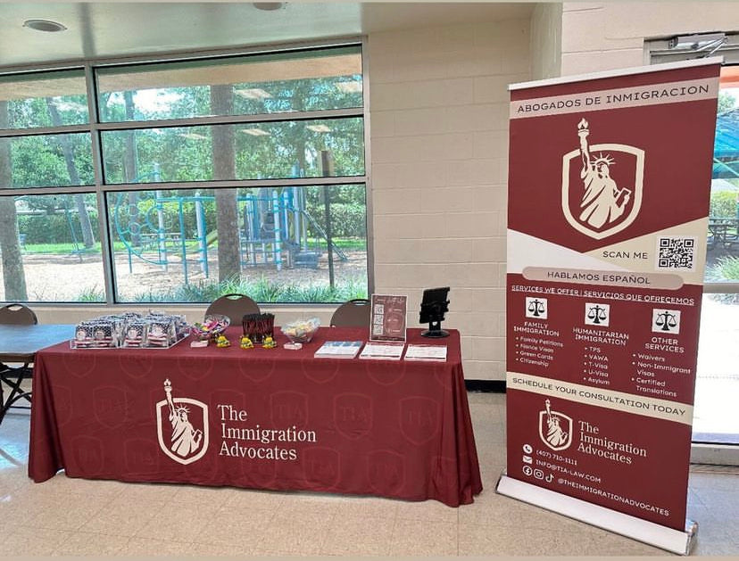 A promotional booth for The Immigration Advocates set up in a large indoor space with a table covered in a dark red tablecloth and a matching vertical banner stand. The branding features a white shield with the Statue of Liberty and the text 'The Immigration Advocates'.