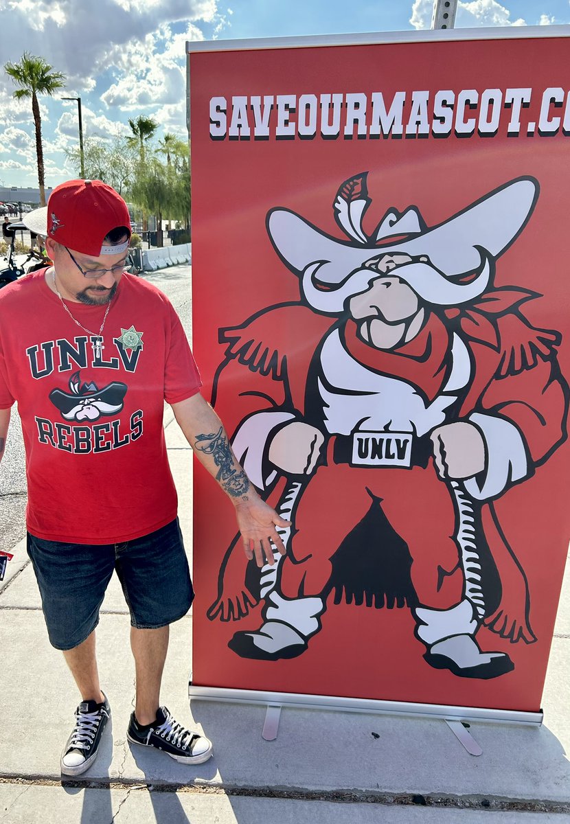A man wearing a red UNLV Rebels t-shirt and shorts stands next to a large red promotional banner featuring the UNLV mascot in a cowboy outfit with the text 'SAVEOURMASCOT.COM'.
