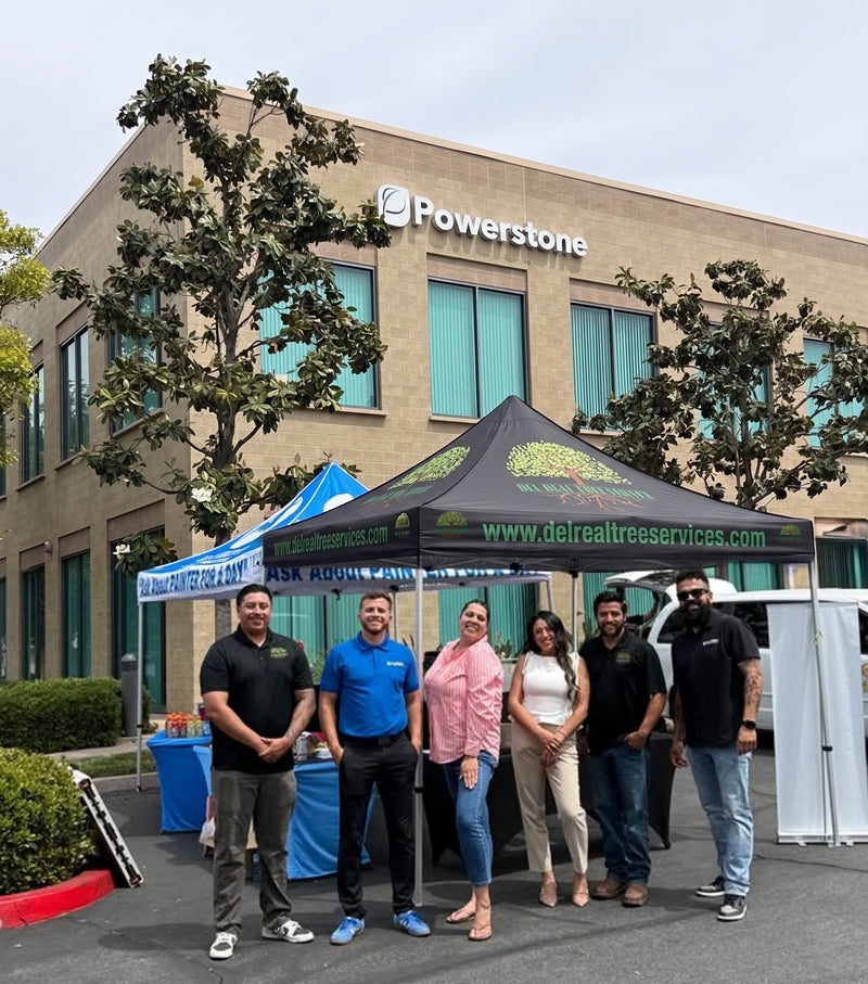 Seven people stand together in front of a Powerstone office building, with a black pop-up canopy tent branded delrealtreeservices.com and a blue table setup in the foreground.