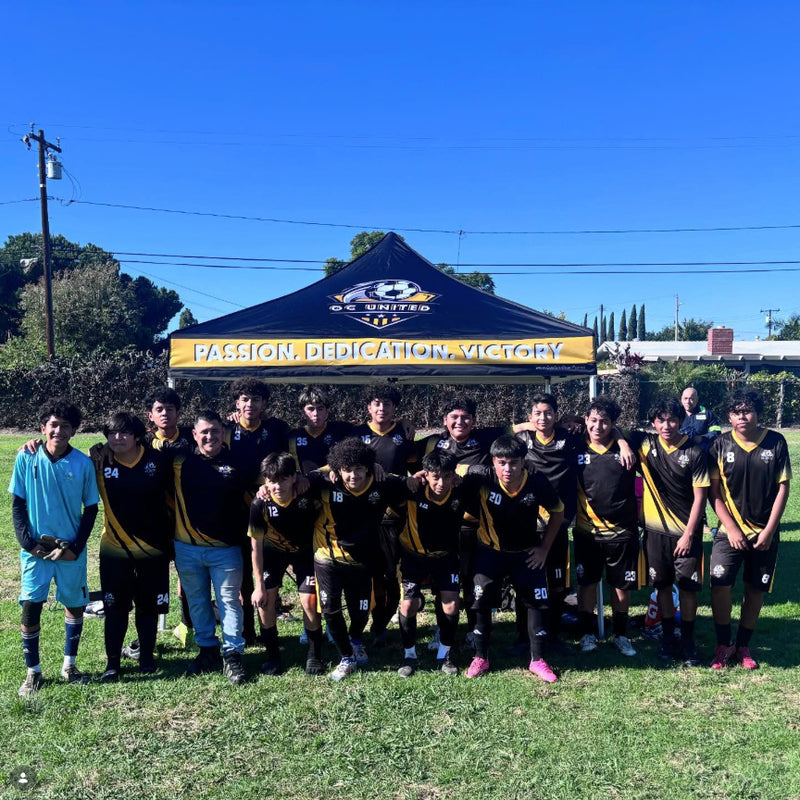 A group of soccer players and coaches posing outdoors in front of a black and yellow team tent on a grassy field under a clear blue sky.