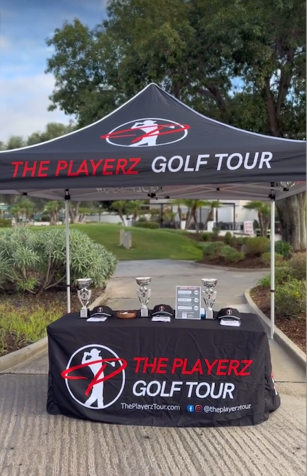 A branded pop-up canopy tent and matching table cover for The Playerz Golf Tour, set up outdoors on a paved path with trees and grass in the background. The canopy is black with red and white branding, and the table is covered in a black cloth with the same logo and text.