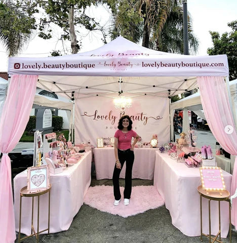 A woman stands behind a pink and white pop-up canopy boutique stall outdoors, with display tables, pink draped curtains, and the brand name Lovely Beauty Boutique printed on the canopy and back wall.