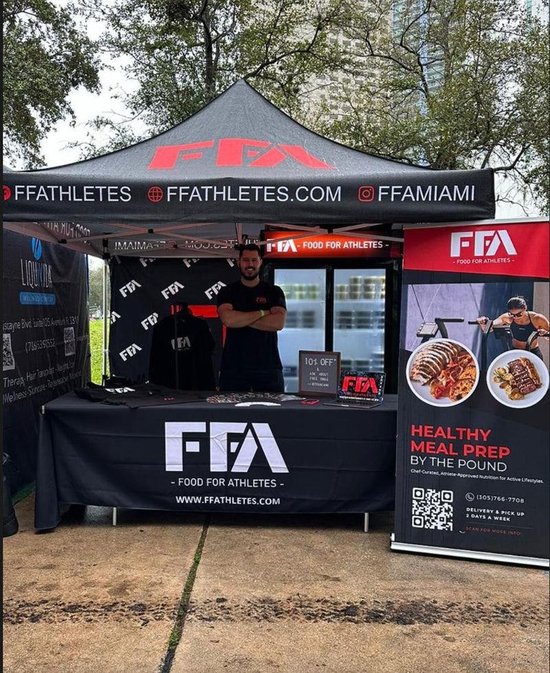 An outdoor promotional booth for FF Athletes / Food For Athletes under a black canopy tent with red and white branding. A man stands behind the table, and printed banners and a sign are displayed on the front and to the right.