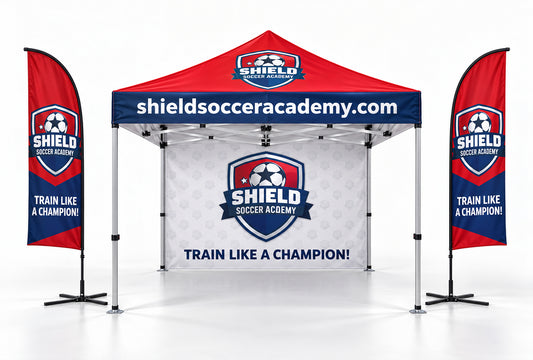 Custom branded soccer academy pop-up canopy tent with two matching vertical feather flags on a light gray background. The canopy and flags feature a red top, dark blue lower band, white center panel, and shield logo for Shield Soccer Academy with the text 'shieldsocceracademy.