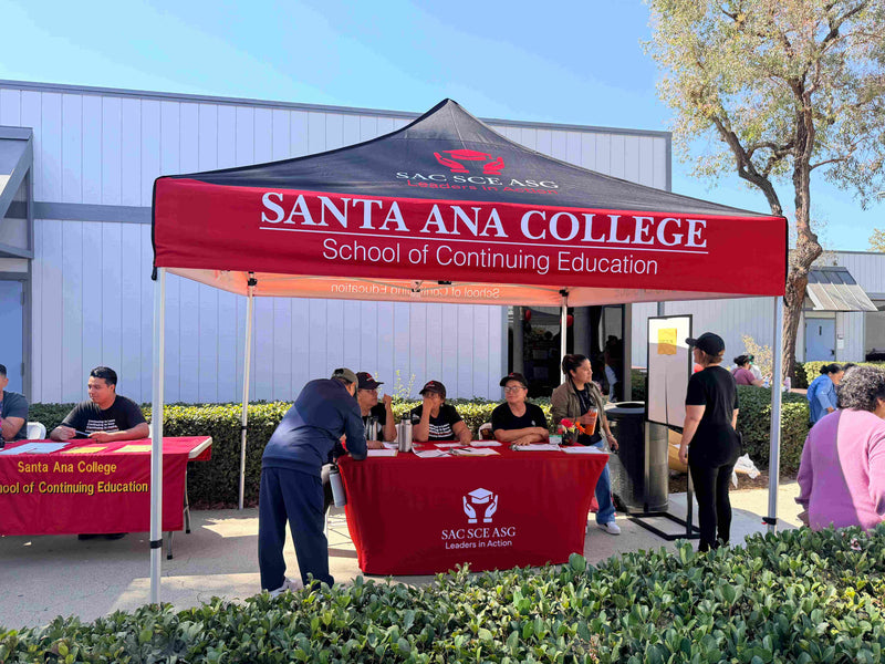An outdoor event booth for Santa Ana College School of Continuing Education under a red and black canopy tent, with people registering or interviewing behind and beside the tables.