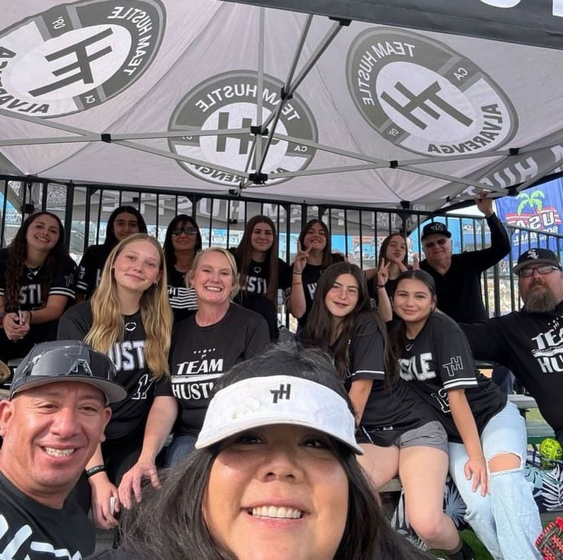 A group of people posing together under a canopy tent with repeated Team Hustle University logos. Most are wearing black Team Hustle shirts, and the scene appears to be a casual outdoor event or game day setting.