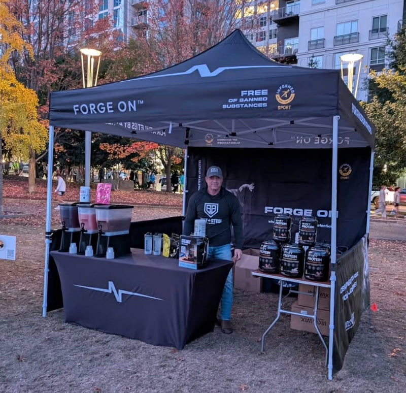 A black branded pop-up canopy tent set up outdoors in a park-like area, with a person standing behind a table inside the tent. The canopy and tablecloth show the brand name Forge On, along with supplement containers displayed on the table and inside the tent.