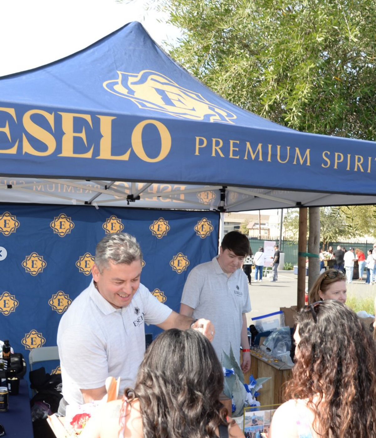A blue Eselo Premium Spirits pop-up canopy tent with yellow lettering, set up at an outdoor event. Several people are gathered around a table under the canopy, with trees and a sunny outdoor background visible.