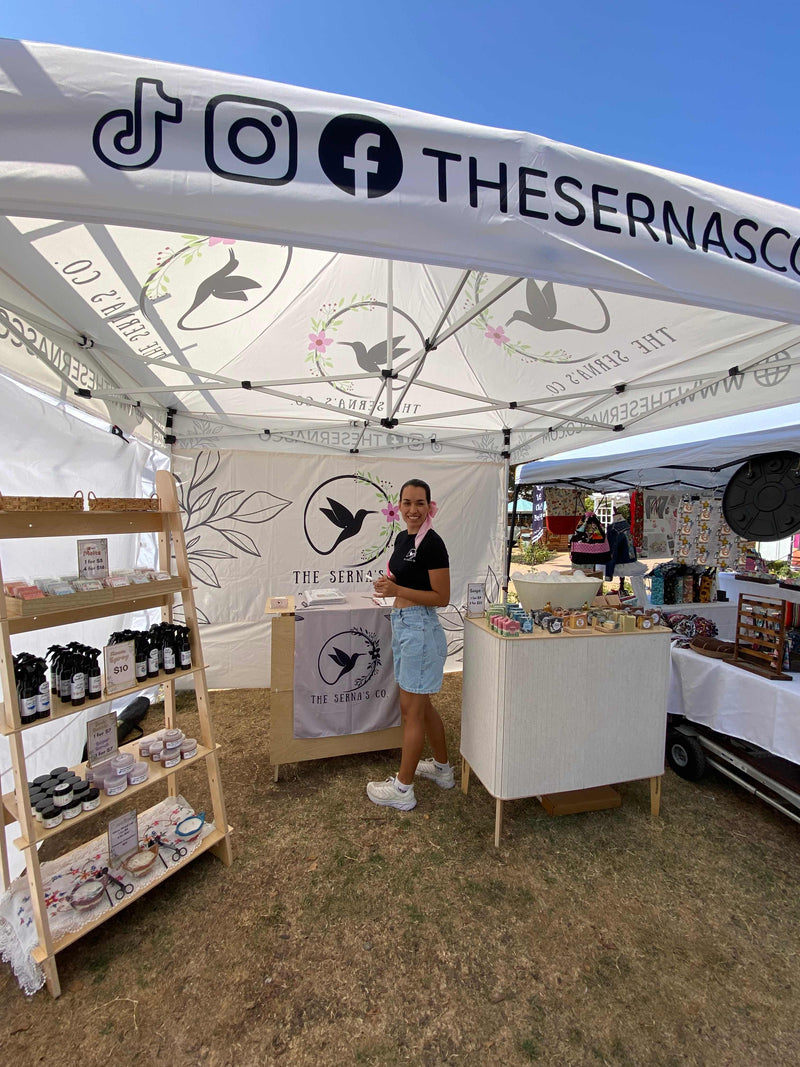 An outdoor market booth under a white canopy tent branded 'THE SERNASCOTT' with bird illustrations. A woman stands behind a counter, with shelves of packaged goods on the left and display tables on the right.