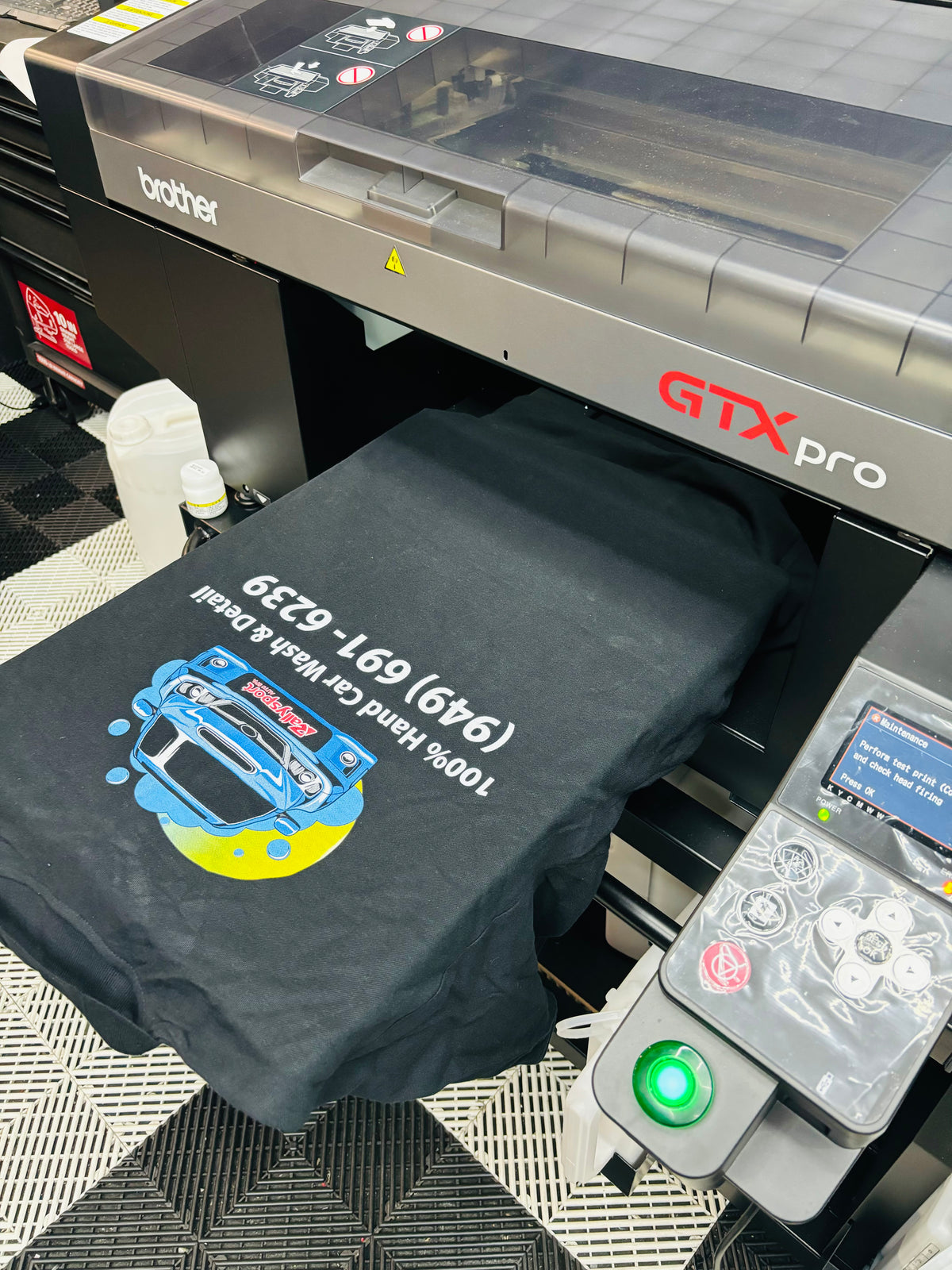 A black t-shirt with a custom car graphic and phone number printed on it, emerging from a Brother GTX Pro UV printer. The t-shirt features a blue and yellow car illustration with white text reading '100% Hand Carved Design'.